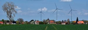 Das Bild zeigt eine Wohnsiedlung an einem Feldrand mit Windkrafträdern im Hintergrund und dient als Beitragsbild zum Blogbeitrag von Janine Wilke Dombert Rechtsanwälte Potsdam und Düsseldorf.